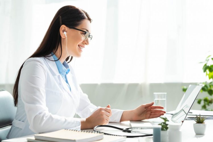 positive-doctor-in-earphones-having-online-consultation-with-patient-on-laptop-in-clinic-office